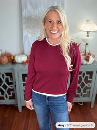 A woman wearing a burgundy crewneck sweater with white trim at the neckline and cuffs, paired with blue jeans. The sweater features a classic, comfortable fit perfect for casual wear.