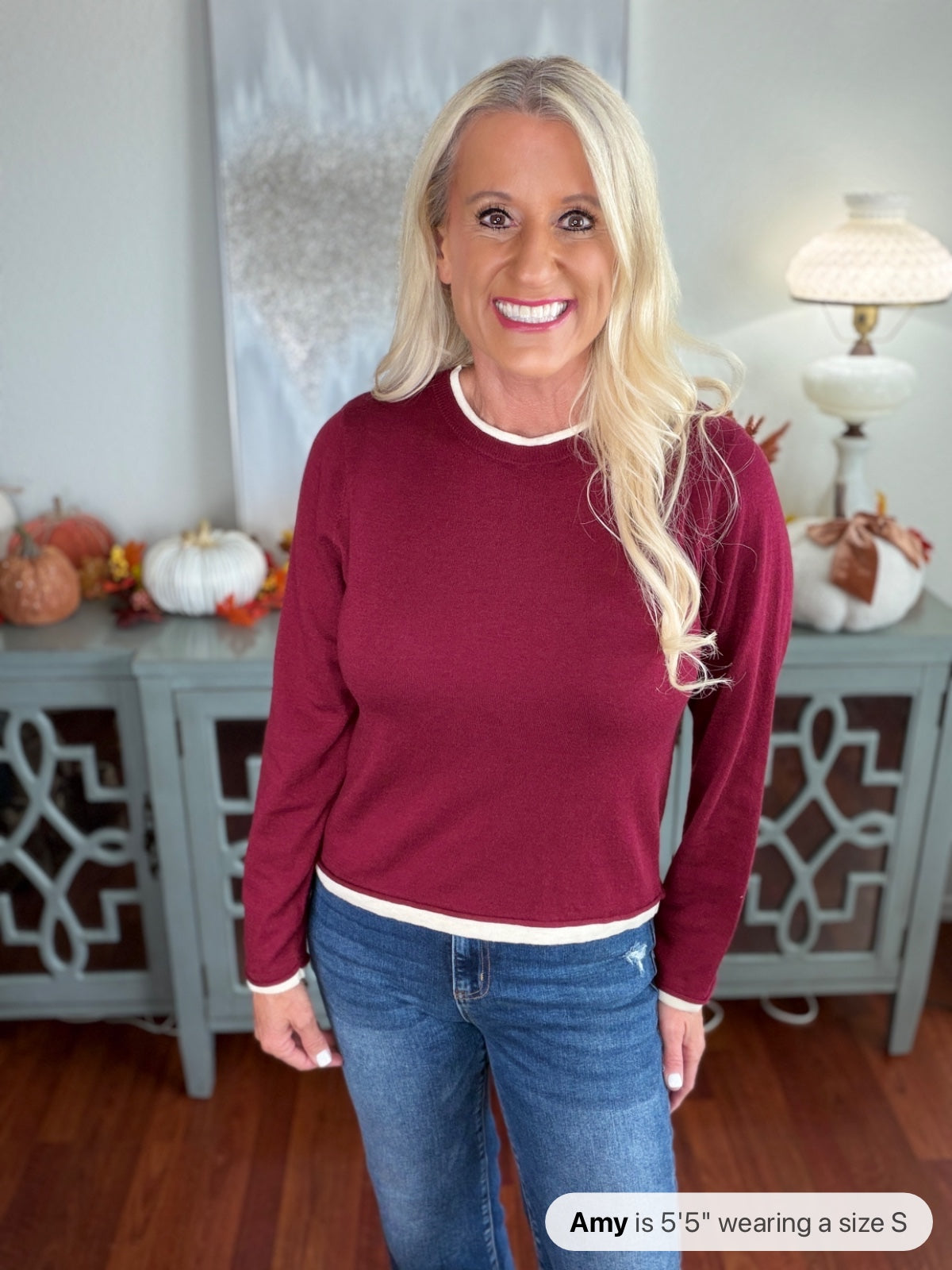 A woman wearing a burgundy crewneck sweater with white trim at the neckline and cuffs, paired with blue jeans. The sweater features a classic, comfortable fit perfect for casual wear.