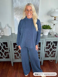 A woman wears a two-piece blue ribbed knit set consisting of a long-sleeve crewneck sweater and matching wide-leg pants, styled with white sneakers.