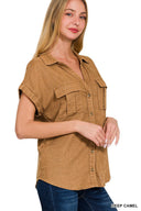 A woman wearing a deep camel-colored short-sleeve button-up shirt with chest pockets and rolled sleeves, paired with dark blue jeans.