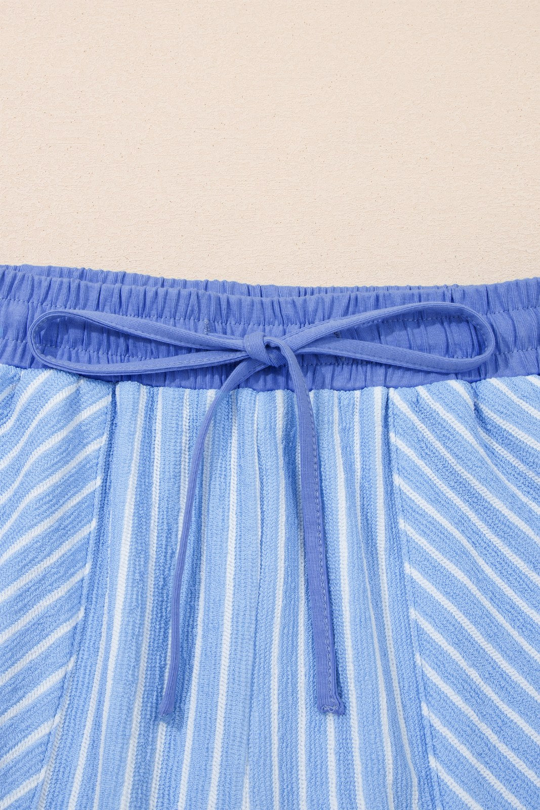 Close-up of blue striped swim shorts with a solid blue drawstring waistband tied in a bow knot. The fabric features vertical white and blue stripes throughout.
