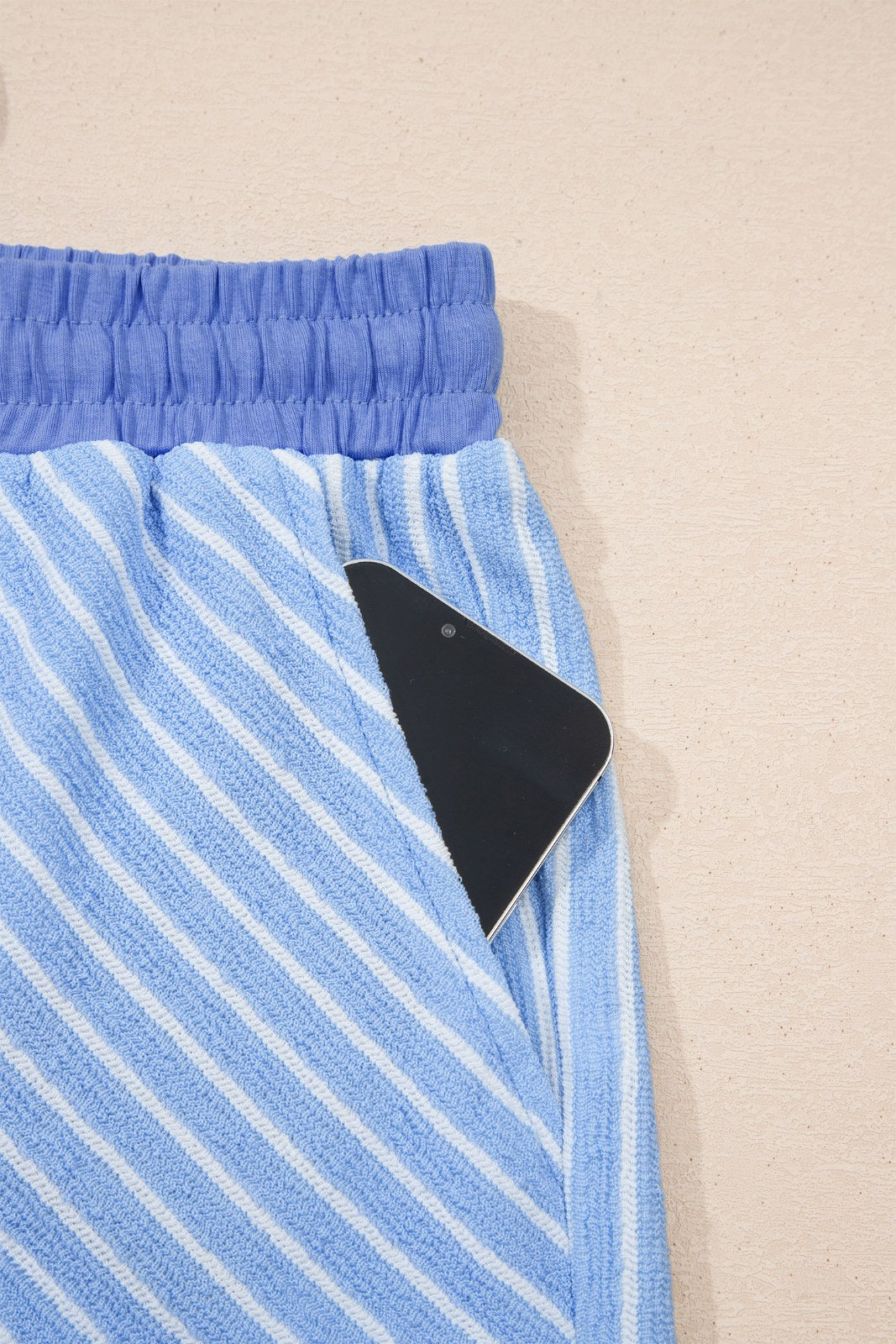 Blue and white striped shorts with an elastic waistband and a black item partially visible in the front pocket, displayed flat against a beige background.
