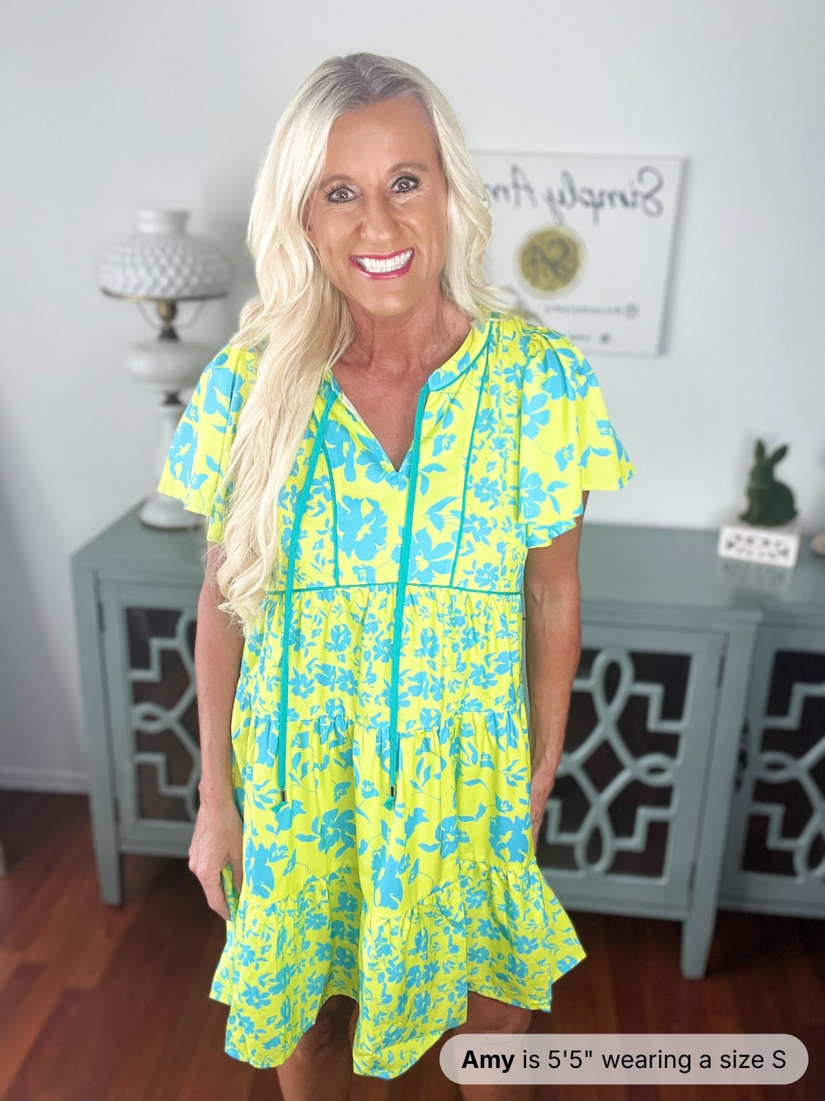 A woman wearing a bright yellow and turquoise floral print short-sleeve dress with a V-neckline and drawstring waist detail, styled with long blonde hair and a bright smile.
