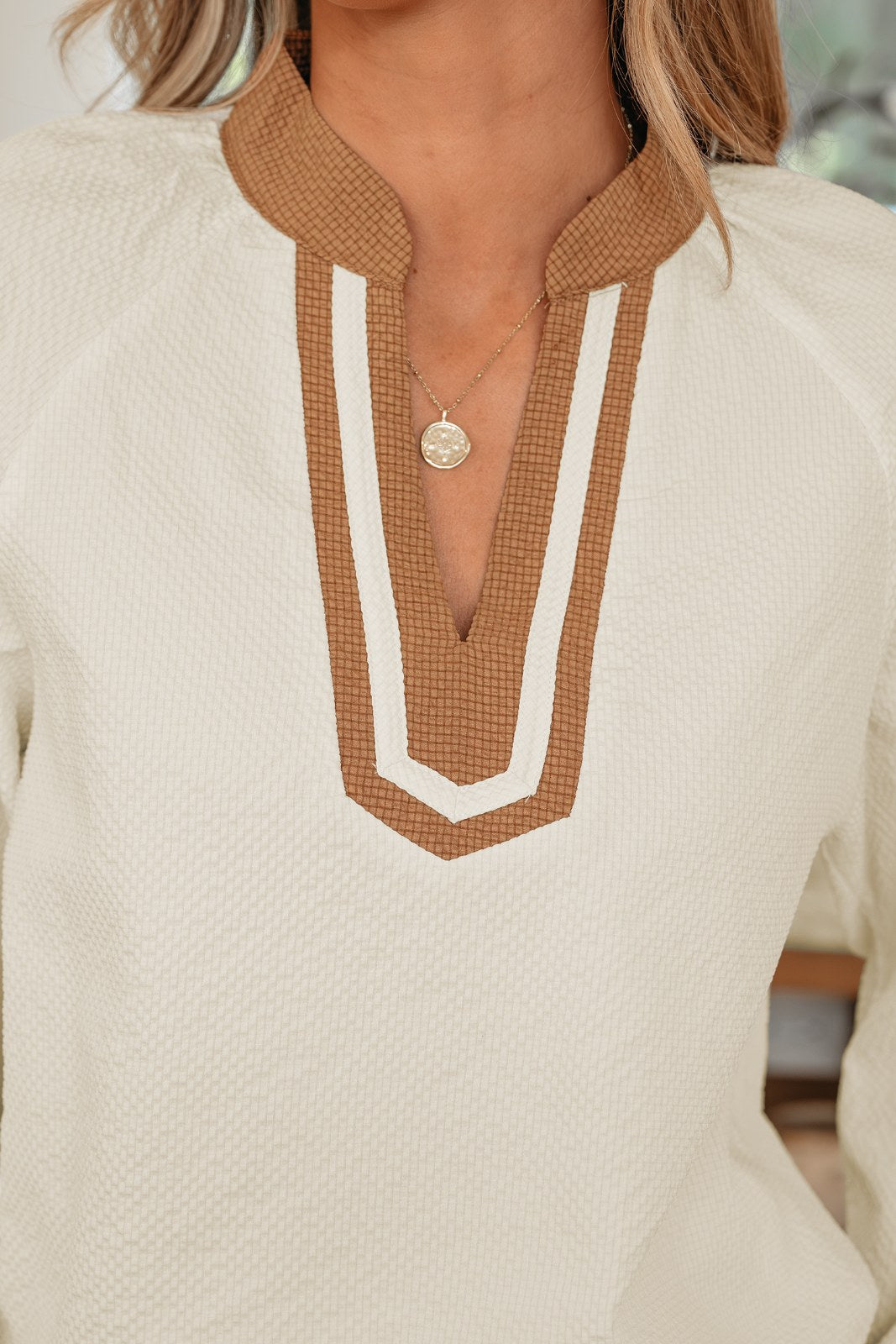 A cream-colored waffle knit top featuring a brown and white geometric patterned collar with a V-neckline, paired with a delicate silver pendant necklace.
