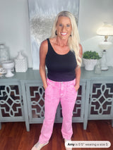 Woman wearing pink denim jeans with a black sleeveless tank top, standing in front of a gray decorative cabinet in a home setting.