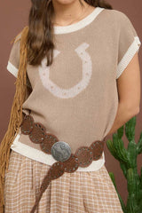 Woman wearing a beige knit top with a horse shoe design, plaid skirt, and brown belt against a brown background.