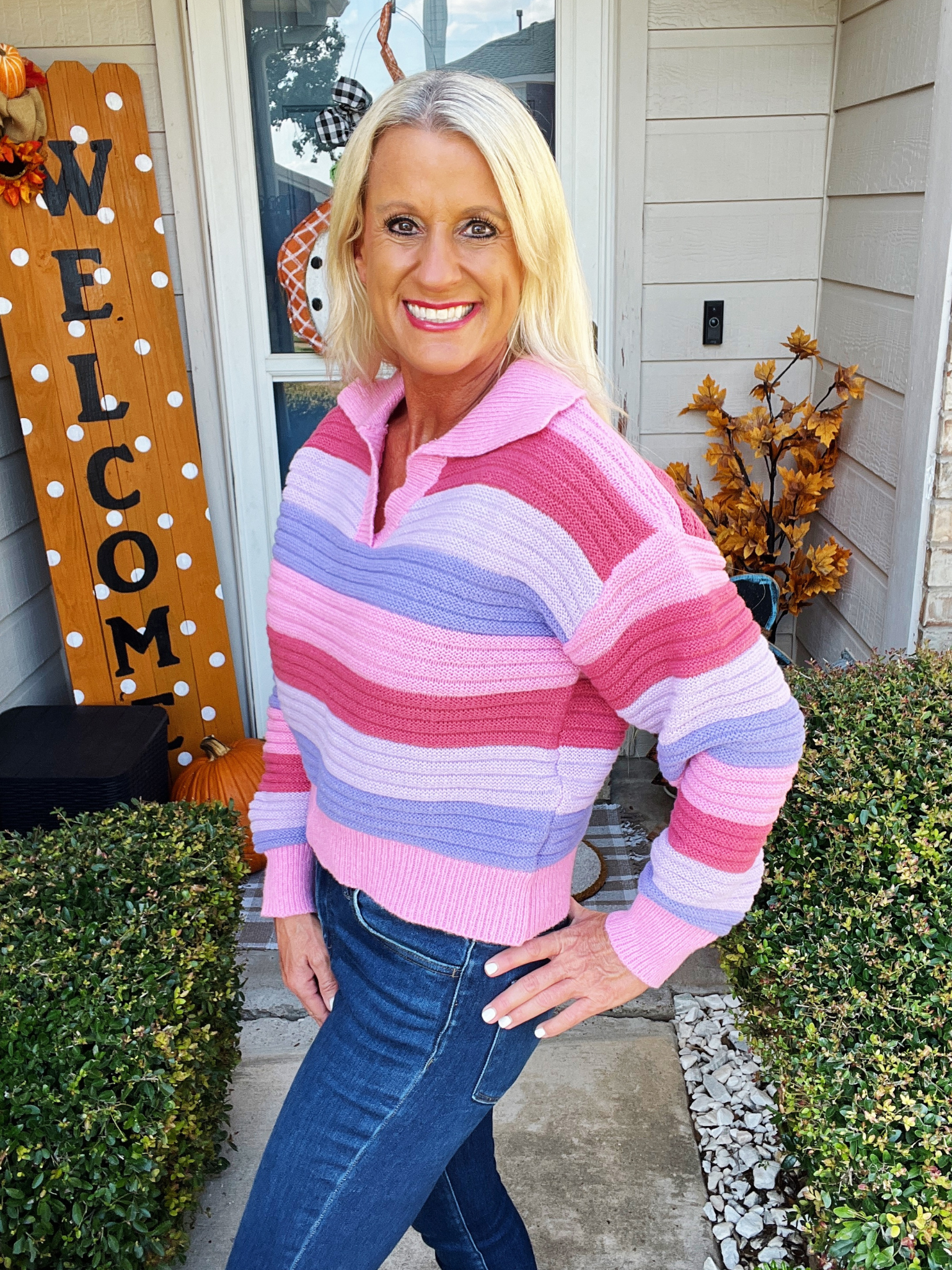 Woman wearing a colorful striped sweater standing in front of a house with 'Welcome' sign and autumn decorations.